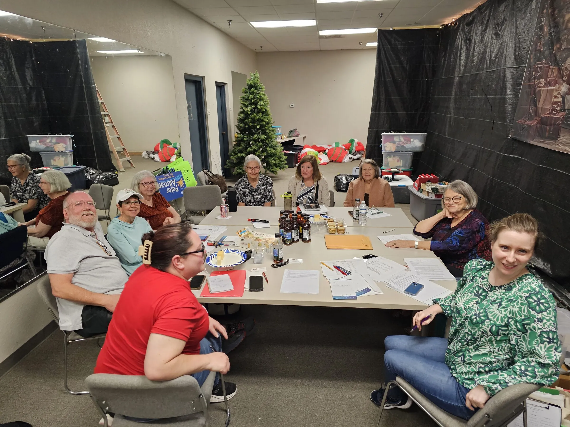 Community members smile around a long table covered with papers and jars at an indoor Brainstorms gathering