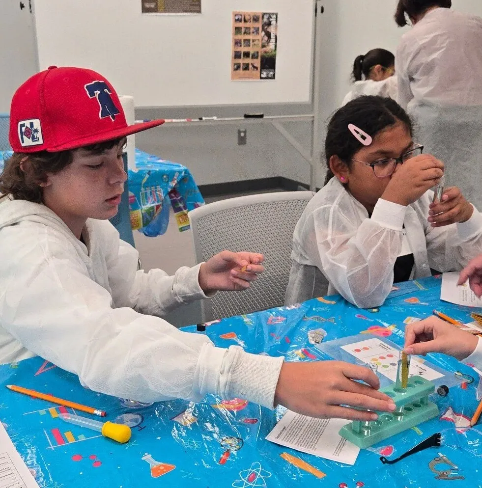 Children in lab coats work side by side on an experiment at a Science Saturday table