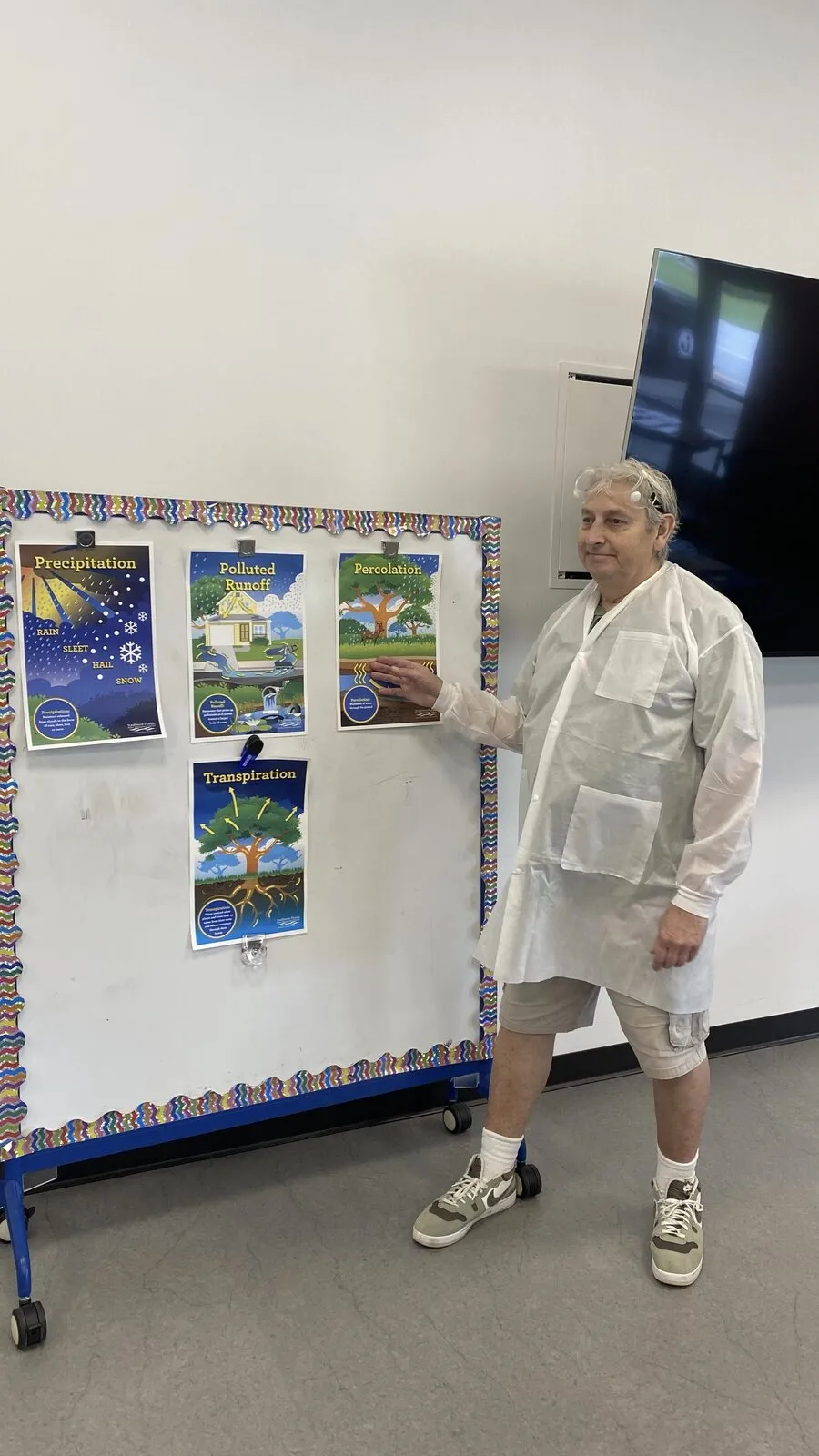 A Brainstorms educator in a lab coat points to a water cycle poster on a display board